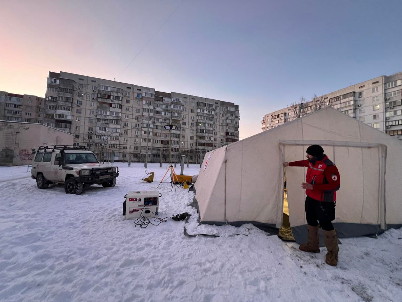 En ung mann i Røde Kors-uniform står utenfor et stort, lyst telt på en snødekt plass, med en firehjulstrekker og boligblokker i bakgrunnen