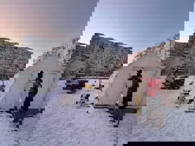 A person in a red jacket stands outside a large tent in a snowy area, with an off-road vehicle and apartment buildings in the background.