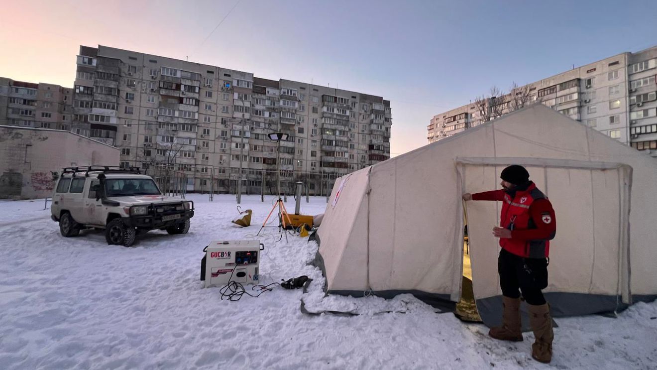 En ung mann i Røde Kors-uniform står utenfor et stort, lyst telt på en snødekt plass, med en firehjulstrekker og boligblokker i bakgrunnen