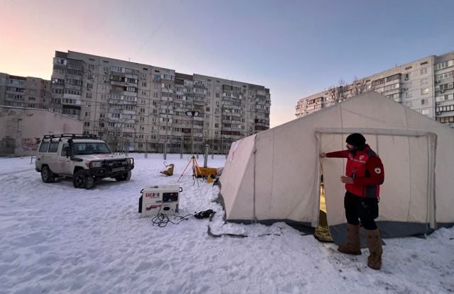 A person in a red jacket stands outside a large tent in a snowy area, with an off-road vehicle and apartment buildings in the background.