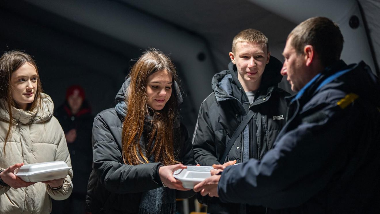 Several young people in jackets receive food containers from a person in uniform inside a tent. Copy response