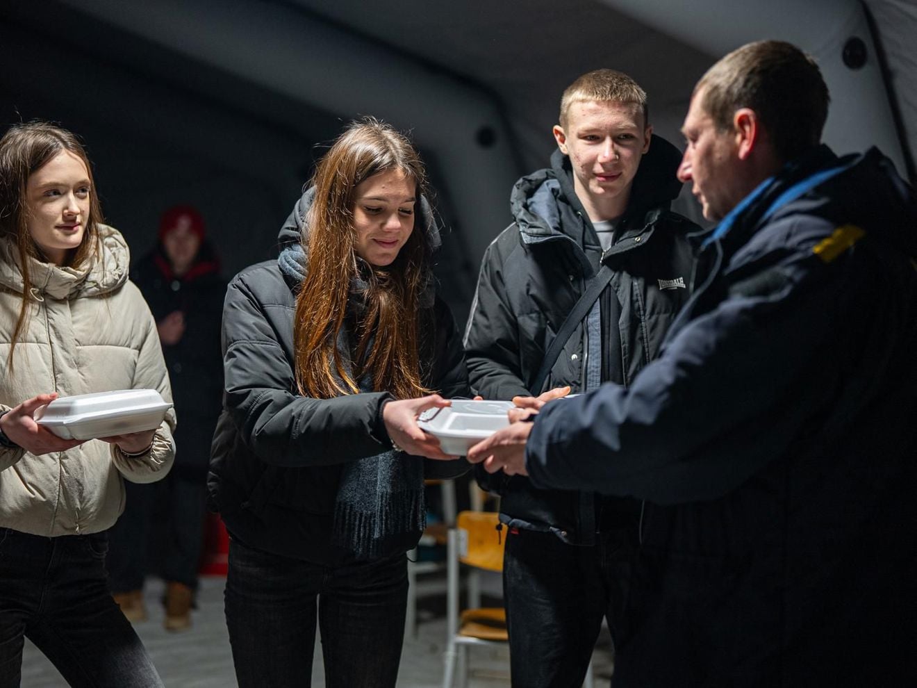 Several young people in jackets receive food containers from a person in uniform inside a tent. Copy response
