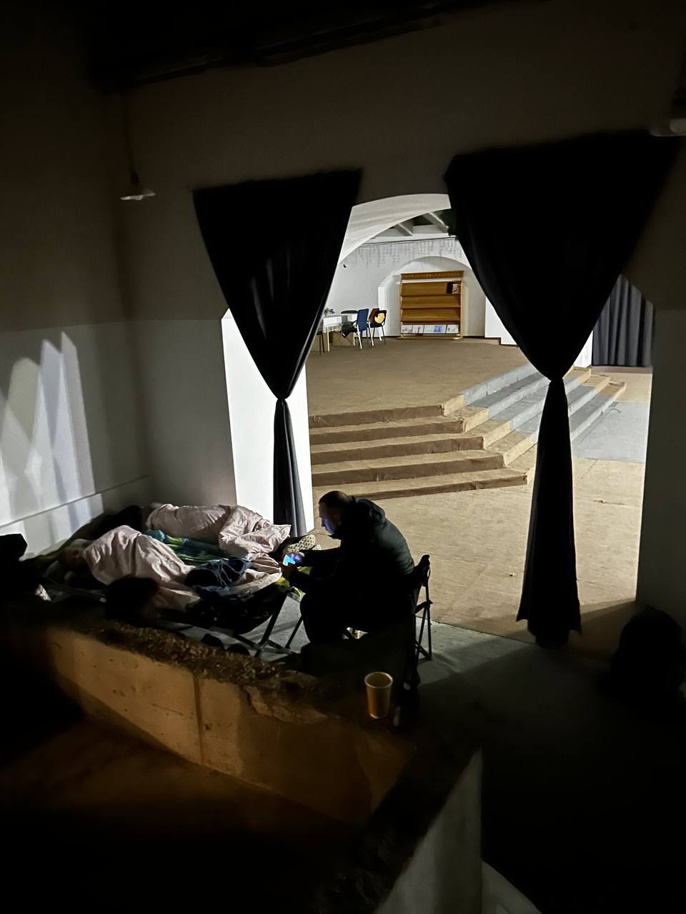 En person sitter ved siden av en feltseng med en sovende under tepper i et mørkt rom, mens et opplyst rom med stoler og hyller vises i bakgrunnen.
