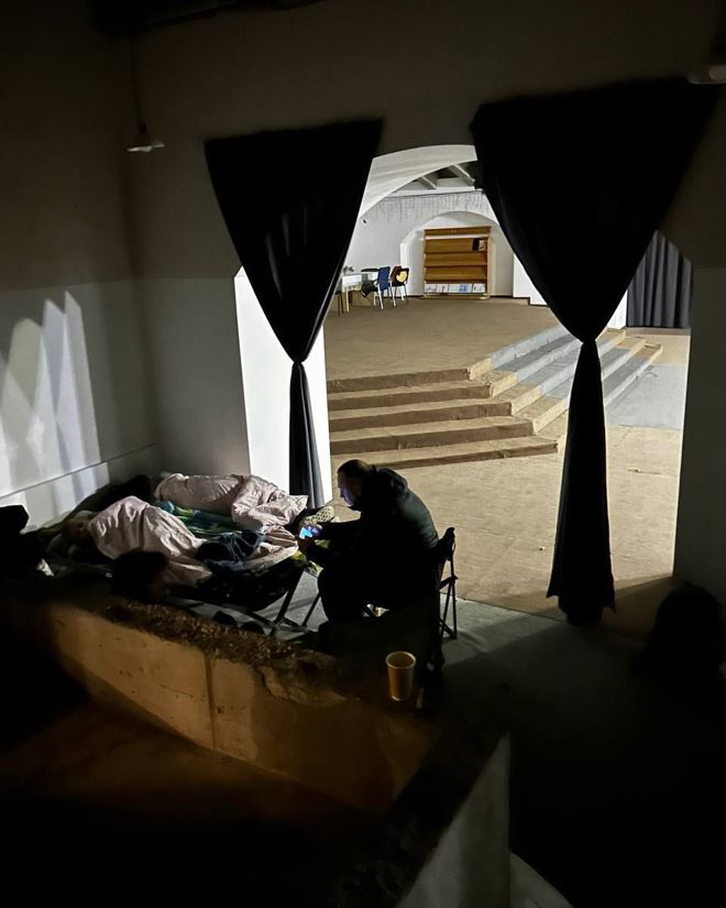 En person sitter ved siden av en feltseng med en sovende under tepper i et mørkt rom, mens et opplyst rom med stoler og hyller vises i bakgrunnen.