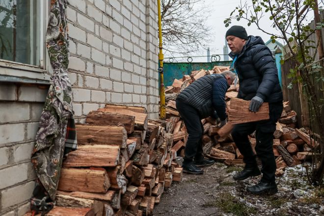 Two adult men are stacking firewood along the side of a brick house.