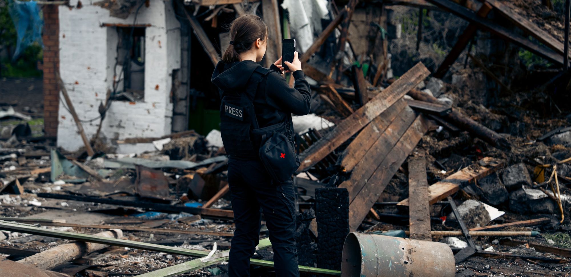 The image shows a person wearing a black press vest standing in front of a destroyed building and large amounts of debris. The individual is holding up a mobile phone or a camera, documenting the destruction.