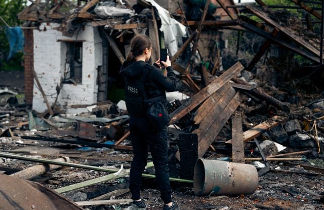The image shows a person wearing a black press vest standing in front of a destroyed building and large amounts of debris. The individual is holding up a mobile phone or a camera, documenting the destruction.