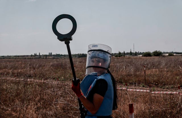 The image depicts an individual wearing protective gear, including a blue vest and a face shield, standing in a dry, open landscape. The person is holding a tool that resembles a metal detector, suggesting they are engaged in work related to safety or demining.