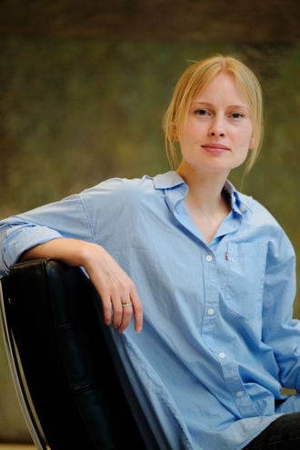 A woman is sitting in a black leather chair, wearing a light blue button-up shirt with rolled-up sleeves. The background is softly blurred in earthy tones, giving the image a calm and relaxed atmosphere. The individual has blond hair tied back and wears a simple ring on one finger.