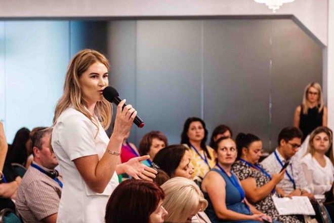 A woman in a white outfit is speaking into a microphone in front of a seated group at a conference or seminar. The audience appears attentive, with some people holding notebooks and wearing name tags. The setting is professional and focused.