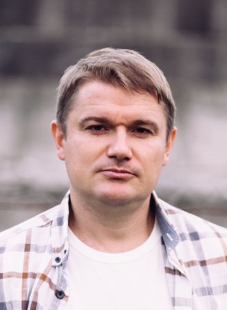 og på engelsk - skriv mann The image shows a man with short light brown hair, wearing a white t-shirt and an open, light-colored plaid shirt. He is outdoors, and the background is softly blurred and grayish, giving the photo a neutral look. The picture is taken from the chest up.