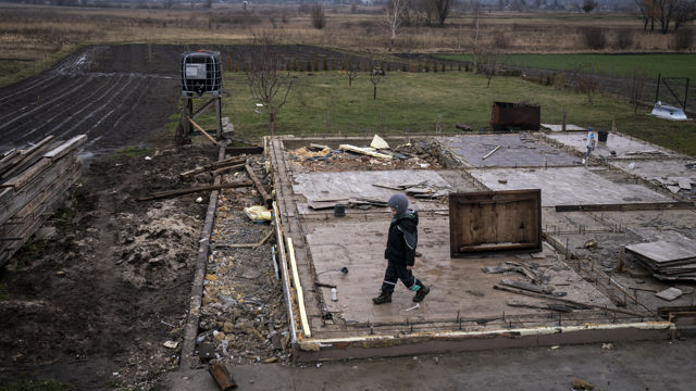 The photo depicts a young child walking across the bare foundation of a demolished house, surrounded by scattered debris and wooden planks. The child is warmly dressed, suggesting cold weather. The background features a rural landscape with fields, patches of grass, and distant trees under cloudy skies, creating a desolate atmosphere.