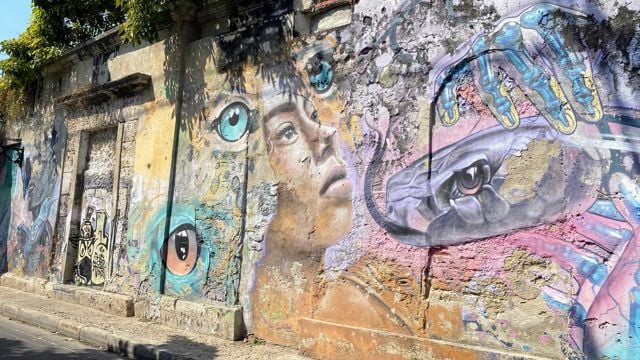 The image shows an old, weathered outdoor wall covered in colorful street art. In the center, there’s a realistic painting of a young person’s face looking upward, surrounded by several large, expressive eyes in different colors. To the right, a large hand with blue and yellow fingers is holding a snake, whose head is painted with a striking, intense eye. The wall’s surface is rough and peeling, with cracks and patches that give the artwork a gritty texture. Sunlight and shadows from nearby trees fall across the top of the wall, adding to the lively and imaginative atmosphere.