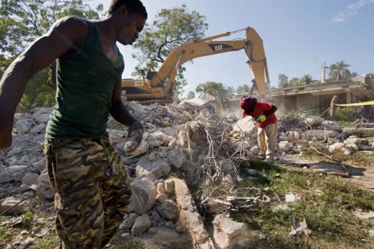 Mann går i en ruin. Gravemaskin i bakgrunnen. Haiti.