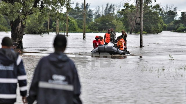 Several people wearing life jackets are being transported in an inflatable boat through a flooded area, while two people stand in the water in the foreground.