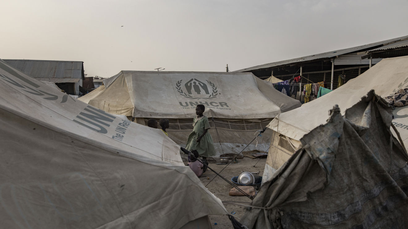 En kvinne og et barn står mellom UNHCR-telt i en flyktningleir, med klær som henger til tørk i bakgrunnen.