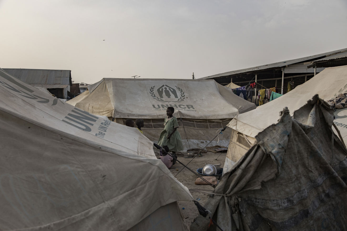 En kvinne og et barn står mellom UNHCR-telt i en flyktningleir, med klær som henger til tørk i bakgrunnen.