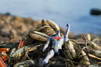 A broken superhero action figure lies among fishing nets and rocks on the shore.