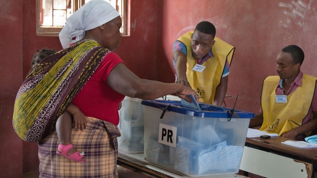 The image depicts a voting scene inside a room with red walls. A person wearing a headscarf and a red shirt is casting a ballot into a transparent box labeled "PR." They have a child secured on their back with a patterned cloth. Two officials wearing yellow vests with name tags are seated at a table, overseeing the voting process.