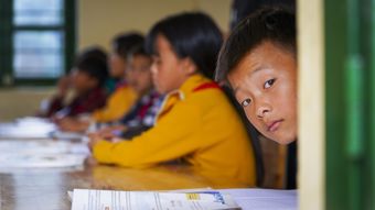 Several young children are sitting at a classroom table with papers and books in front of them.