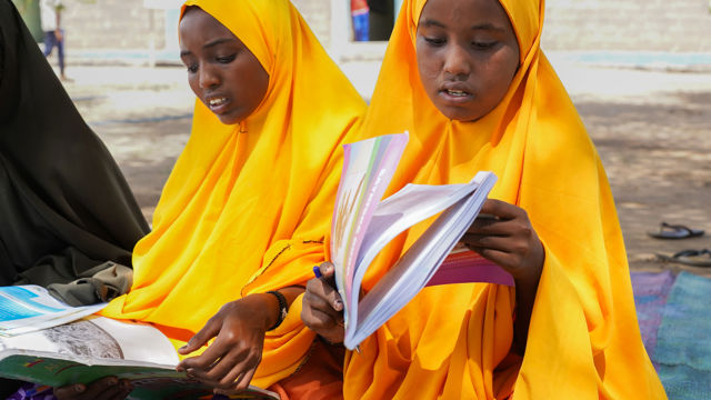 Two girls in yellow hijabs sit outdoors reading schoolbooks.