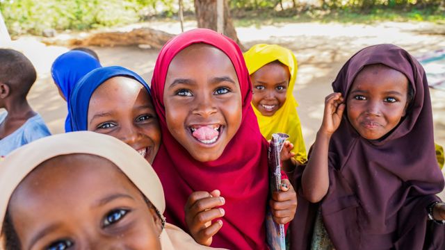 A group of young girls are standing together outdoors, wearing colourful headscarves and brightly coloured clothing.