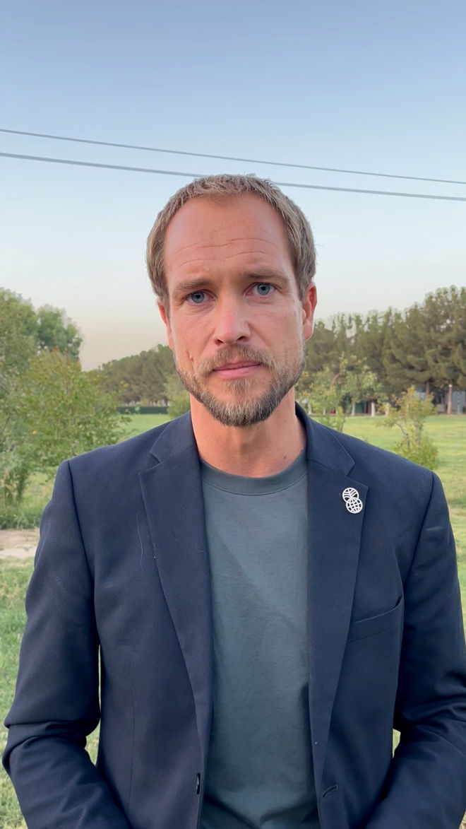 En ung mann med kort hår og skjegg står utendørs med grønn vegetasjon og blå himmel i bakgrunnen.